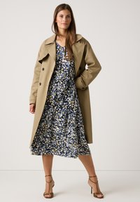 Beiger Trenchcoat über einem dunkelblauen Blumenkleid mit gelben Akzenten. Das Kleid hat eine fließende Silhouette und einen knielangen Saum, kombiniert mit Riemchen-Heels.
