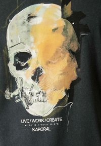 Tissu noir avec un design graphique d'un crâne agrémenté d'accents abstraits jaunes et verts. Le texte en dessous indique "VIVRE/TRAVAILLER/CRÉER KAPORAL."