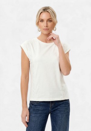 Blondynka ubrana w biały top z krótkimi rękawkami i koronkowymi detalami oraz niebieskie jeansy, pozująca z ręką przy brodzie na białym tle.