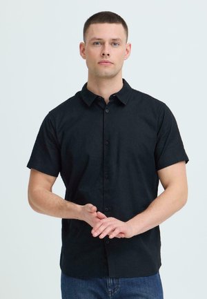 Młody mężczyzna z krótkimi włosami, ubrany w czarną koszulę na krótki rękaw i niebieskie dżinsy, zgromił dłonie przed białym tłem.