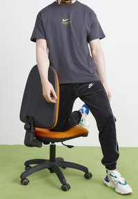 Černá ergonomická kancelářská židle s oranžovým polštářem na sedadle, osoba oblečená v tmavě šedém tričku Nike a černých kalhotách, barevné tenisky.