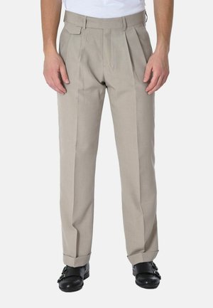 Uomo con pantaloni beige plissettati con orli risvoltati e scarpe nere con fibbia, in piedi davanti a uno sfondo bianco uniforme.