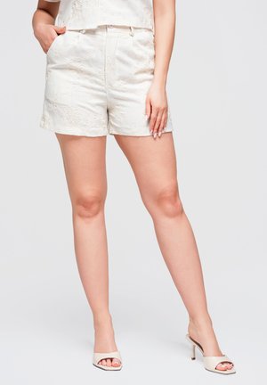 Kvinde iført hvide broderede shorts og beige åbne hælede sandaler, står med den ene hånd i lommen og den anden hånd hvilende på låret.