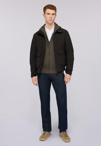 Černá zipová bunda přes olivově zelenou mikinu, spárovaná s tmavě modrými džíny a béžovými teniskami. Obsahuje přední kapsy a jednoduchý design.