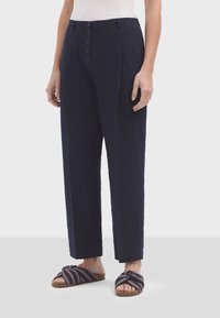 Pantalone širokog kroja u mornarsko plavoj boji s teksturom, visokog struka, s pet dugmadi i prednjim plitkim naborima. Upareni s natikačama s prugicama.