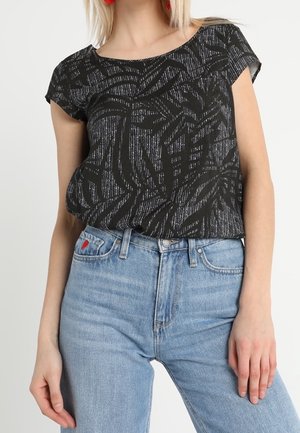 Kobieta nosząca czarny, wzorzysty top z krótkim rękawem, wsunięty w jasnoniebieskie, wysokowaistowe dżinsy z czerwonym haftem serca na kieszeni.