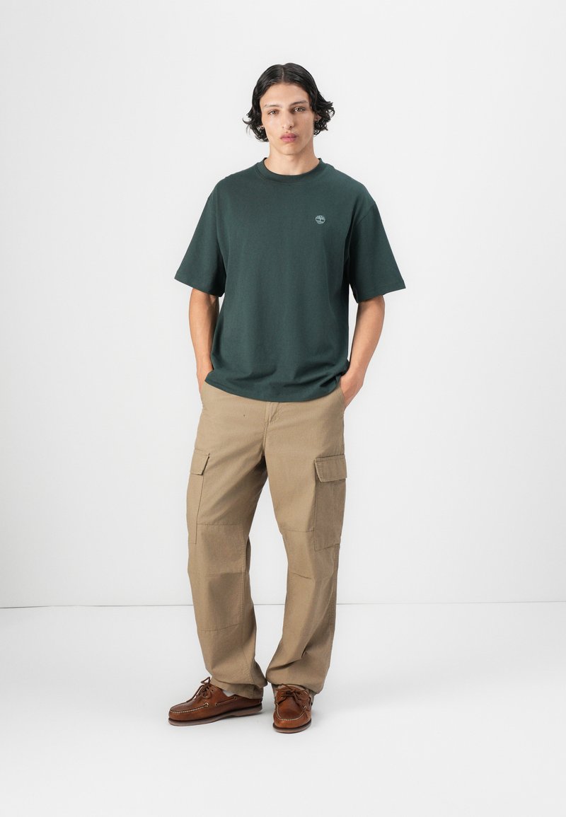 Camiseta de algodón verde oscuro de mangas cortas, combinada con pantalones cargo beige que tienen múltiples bolsillos, y zapatos marrones con cordones.