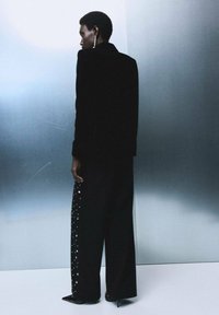 Blazer de terciopelo negro con hombros estructurados, combinado con pantalones de pierna ancha de talle alto en negro, que tienen adornos plateados en los laterales.