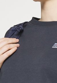 Sudadera azul marino con cuello redondo, tejido texturizado y un pequeño logo geométrico en el pecho. Una mano descansa sobre la costura del hombro.