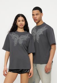 Mujer joven y hombre con camisetas oversized de color gris oscuro a juego, con diseños abstractos simétricos de alas en el pecho, de pie uno al lado del otro.