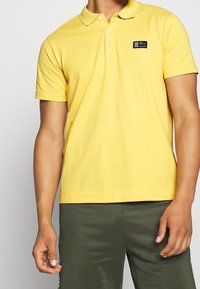 Homme portant un polo à manches courtes jaune avec un patch logo rectangulaire noir et un pantalon vert olive, se tenant contre un fond uni.