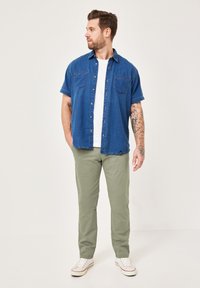 Kurzärmliges Jeanshemd in Dunkelblau mit zwei Brusttaschen, kombiniert mit hellgrünen Hosen und weißen Sneakers. Glatte Textur, lässige Passform.