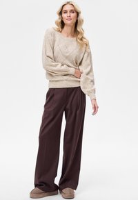 Beżowy dziany sweter z wzorem rombów, z bufiastymi rękawami, w zestawieniu z wysokim stanem, szerokimi, ciemnobrązowymi spodniami oraz beżowymi kapciami.