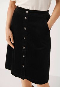 Black corduroy skirt featuring a button front, side pockets, and a slightly flared hem. The texture is ridged, with shiny metallic buttons.