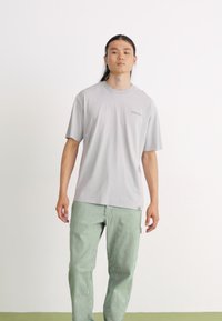 T-shirt cinzento de manga curta com decote redondo e detalhe de logotipo discreto, combinado com calças verdes listradas. Textura de tecido suave.