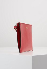 Bolso de mano de cuero rojo con textura lisa, forma rectangular delgada y bordes negros; presenta un pequeño detalle de logo dorado en el frente.