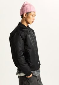 Black bomber jacket with a smooth, shiny finish, ribbed cuffs, and a high collar; worn with a pink knitted beanie.