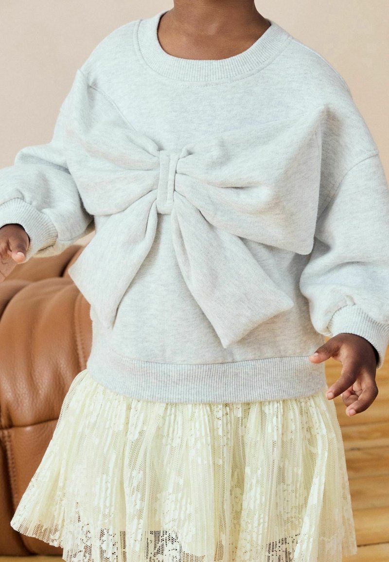Enfant portant un pull gris clair avec un grand nœud et une jupe en dentelle crème, debout à l'intérieur près d'un canapé marron.