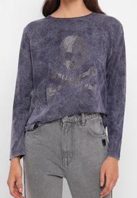 Langes lila Oberteil mit verblasstem Finish, das ein Rhinestone-Schädel-und-Kreuzbones-Design zeigt. Kombiniert mit grauen Jeans.