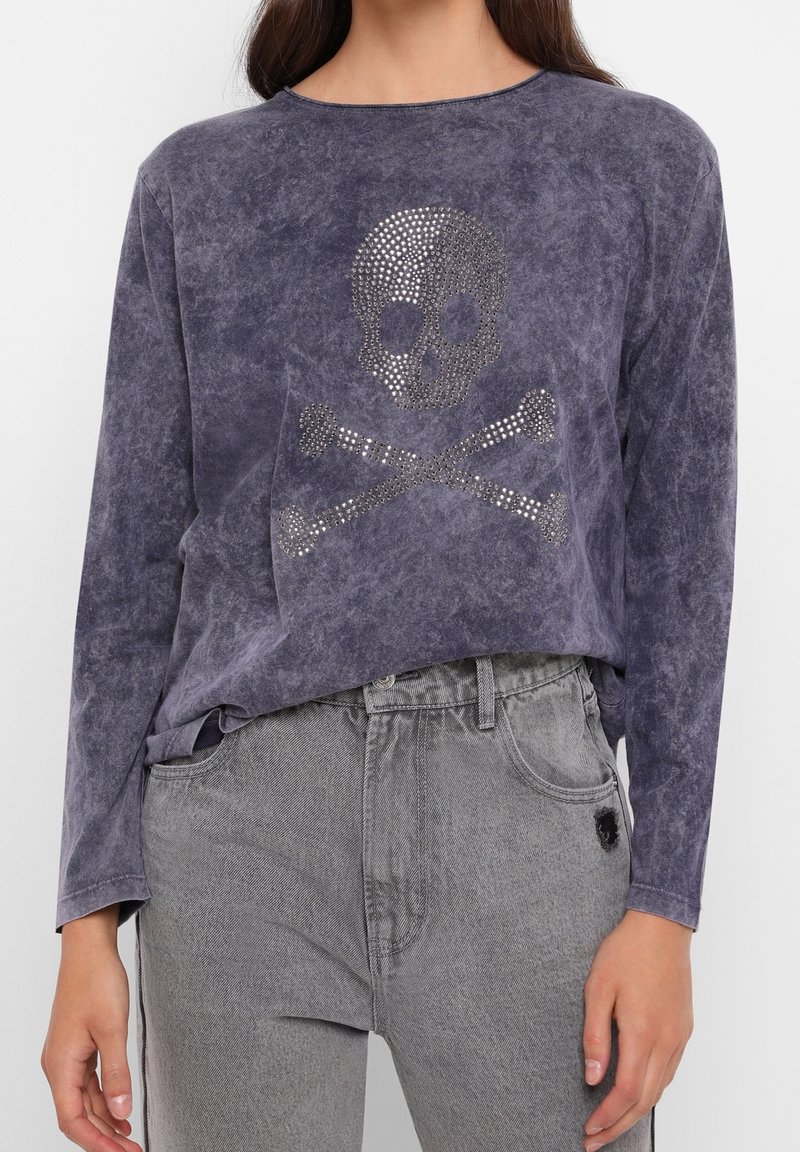 Langes lila Oberteil mit verblasstem Finish, das ein Rhinestone-Schädel-und-Kreuzbones-Design zeigt. Kombiniert mit grauen Jeans.