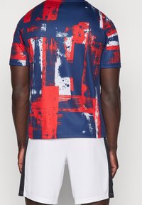 Tričko s krátkým rukávem s vícebarevným abstraktním vzorem malby v červené, modré a bílé barvě. Vyrobeno z hladkého, materiálu odvodu vlhkosti.