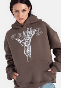 Brązowy hoodie z dużą białą grafiką dłoni. Wzór zawiera kropelki i tekst, ma przednią kieszeń oraz ściągacze na mankietach.