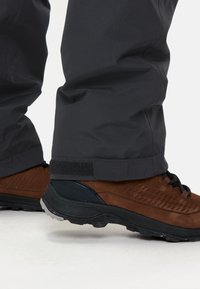 Braune Wanderschuhe mit strukturiertem Wildleder-Obermaterial, schwarzen Akzenten und flexiblen, robusten Sohlen, kombiniert mit schwarzen wasserdichten Hosen mit Klettverschlüssen.