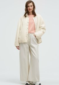 Cream faux fur jacket with snap buttons, worn over a light pink t-shirt and beige wide-leg pants. White sneakers visible.