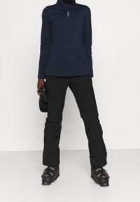 Persona vestita con pantaloni da sci neri e stivali, maglia a maniche lunghe blu navy, tiene dei guanti neri, in piedi davanti a uno sfondo bianco.