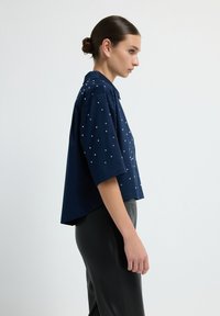 Camicia a maniche corte di colore blu navy con colletto, caratterizzata da decorazioni quadrate bianche in tutto il tessuto. Vestibilità rilassata, design corto e texture liscia.