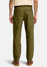 Pantaloni verde oliva con una vestibilità dritta, realizzati in tessuto testurizzato, con due tasche posteriori e un'etichetta con il logo sulla vita. Calzature marroni visibili.