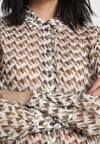 Donna che indossa una camicia beige con bottoni e un motivo ripetuto di teste di cavallo marroni, braccia incrociate e capelli scuri di lunghezza media.