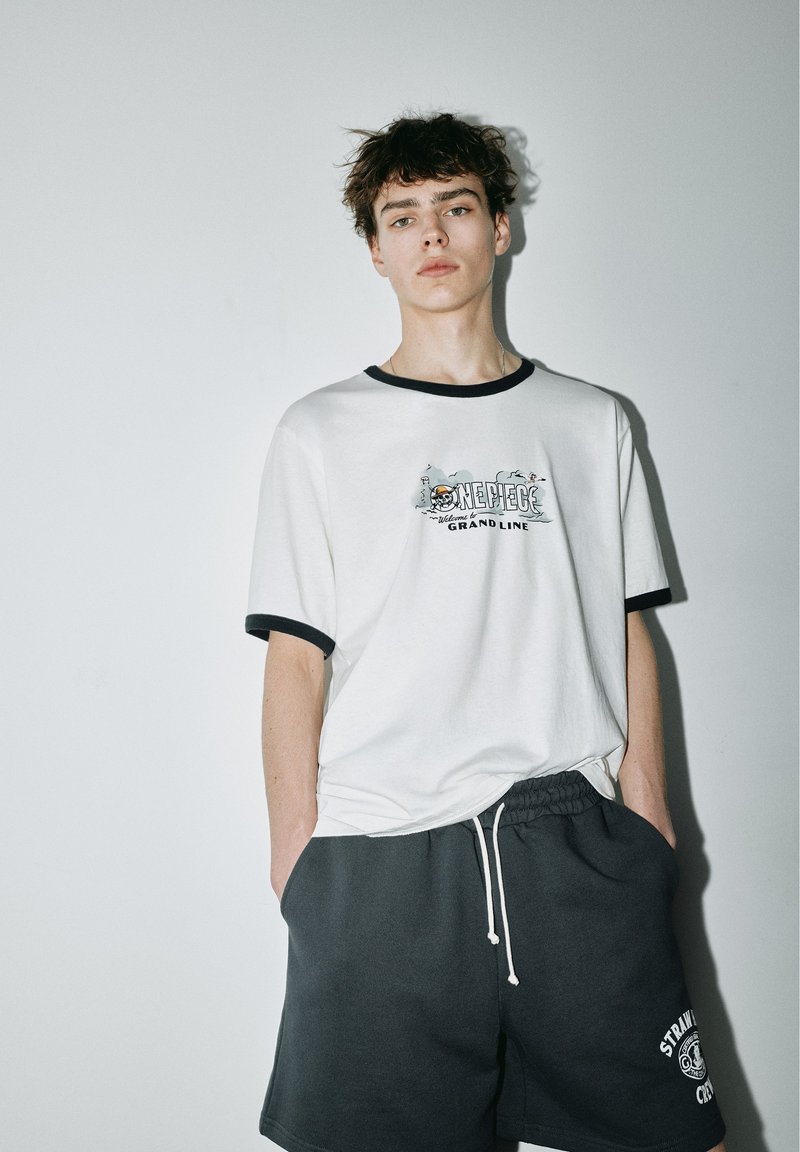 Jeune homme debout contre un mur blanc, portant un t-shirt blanc à motif One Piece et un short gris foncé avec cordon, les mains dans les poches.
