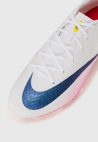 White soccer shoe with a textured finish, featuring a large dark blue Nike swoosh, white laces, and a pink-tinged sole.