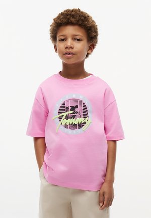 Młody chłopiec z kręconymi włosami, ubrany w różowy t-shirt z nadrukiem i beżowe spodnie, stojący na tle jednolitego białego tła.