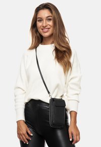 Black leather crossbody bag with a zippered closure and flap, worn on a black leather pant, paired with a white, textured sweater.