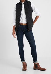 Person iklädd en vit skjorta, svart vadderad väst, mörka slim-fit jeans, brun bälte med guldankarbälte, och bruna ankelstövlar, stående med en hand i fickan.