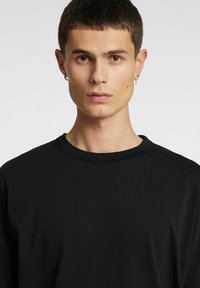 Juoda marškinėliai su apvalia apykakle, lygiu paviršiumi ir trumpomis rankovėmis. Modelis rodo neutralią išraišką ir turi trumpus plaukus.