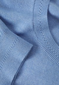 Materialul puloverului tricotat de culoare albastră deschisă are o textură netedă. Dispune de un guler și manșete ribbed, cu cusături decorative subtile pe guler.
