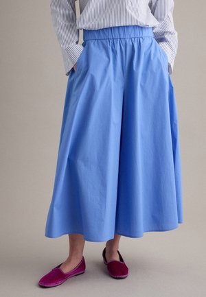 Persona que lleva una falda azul suelta hasta la rodilla, una camiseta de manga larga a rayas y zapatos magenta sin cordones, de pie con las manos en los bolsillos.