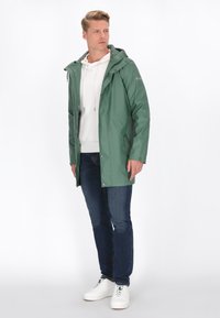 Vízálló zöld dzseki, kapucnival, ezüst színű hardverrel és oldalsó zsebekkel. Fehér kapucnis pulóverrel, kék farmernadrággal és fehér sportcipővel párosítva.