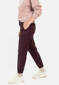 Bordeauxfarbene tapered Hosen mit elastischen Bündchen, aus glattem Stoff. Getragen mit einem hellrosa Pullover und neutralen Turnschuhen. Seitentaschen sichtbar.