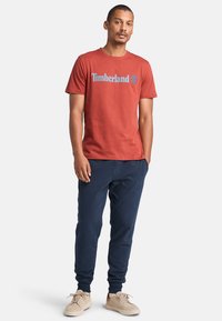 Červené bavlněné tričko s modrým logem "Timberland", doplněné tmavě námořnickými tepláky a světle béžovými teniskami. Jednoduchý, casual design.