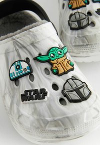 Weiße Clogs mit Marmor-Muster und mehreren Löchern; ausgestattet mit Gummisteckern, die Figuren und Logos aus einem Science-Fiction-Franchise darstellen.