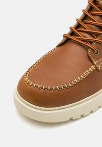 Bota de cuero marrón con una puntera cosida, cordones naranjas y una suela de goma blanca texturizada, que presenta un acabado suave y costuras detalladas.