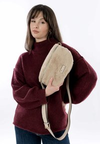 Sac bandoulière en fausse fourrure beige avec une fermeture éclair argentée et une sangle beige, tenu contre un pull bordeaux. La texture semble douce et pelucheuse.
