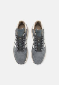 Sneaker in suede grigio con tre strisce bianche, lacci neri e suola in gomma beige. Presentano un collo imbottito e un marchio dorato sulla linguetta.