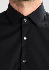 Close-up van de nek van een man die een zwart collshirt draagt met drie zilveren knopen vastgemaakt, met een lichte huidstoppeling zichtbaar.