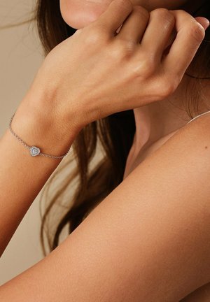 Bracciale in argento con un pendente a forma di cuore, dotato di una pietra centrale circondata da pietre più piccole, indossato su un polso.