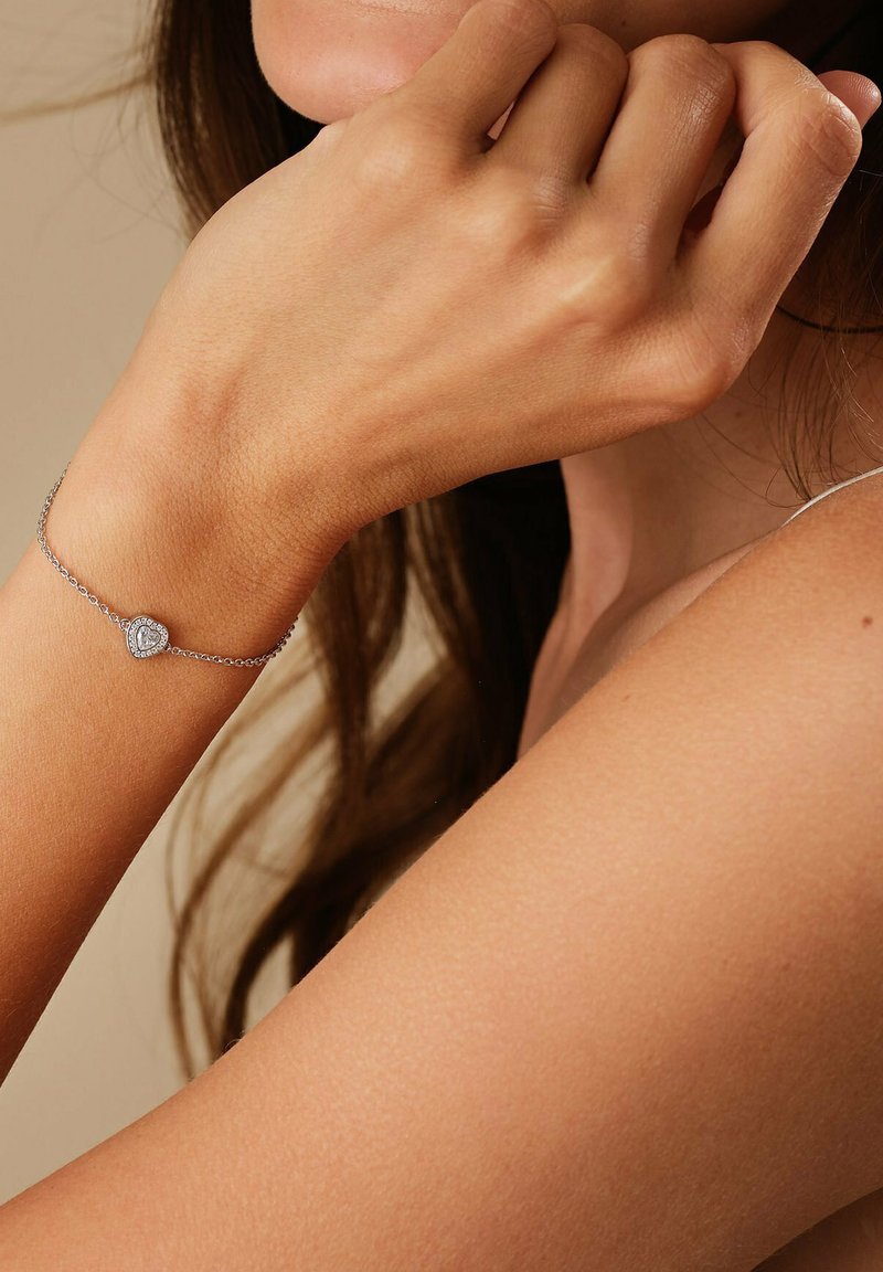 Silbernes Armband mit einem herzförmigen Anhänger, der einen zentralen Edelstein umgeben von kleineren Steinen zeigt, am Handgelenk getragen.